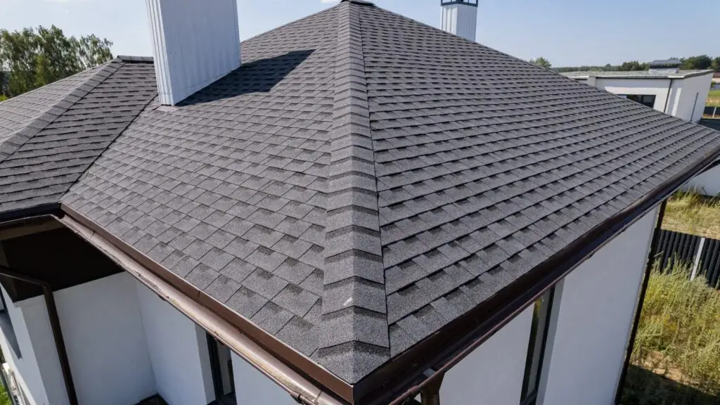 Aerial view of a modern house with a gray asphalt shingle roof. The house has white walls and brown gutters, surrounded by greenery and a clear blue sky in the background. Dominion Roofing | BRAMPTON | BURLINGTON | ETOBICOKE | MARKHAM | MISSISSAUGA | NORTH YORK | OAKVILLE | RICHMOND HILL | Scarborough | TORONTO | VAUGHAN | dominionroofing.com | Phone Number: 416-789-0601
