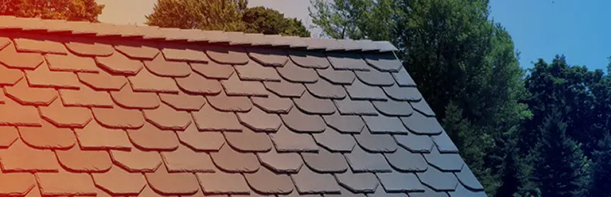 Close-up of a tiled roof with overlapping rectangular shingles, in a gradient light from red to blue on the left. In the background, greenery with tall trees under a clear blue sky. Dominion Roofing | BRAMPTON | BURLINGTON | ETOBICOKE | MARKHAM | MISSISSAUGA | NORTH YORK | OAKVILLE | RICHMOND HILL | Scarborough | TORONTO | VAUGHAN | dominionroofing.com | Phone Number: 416-789-0601