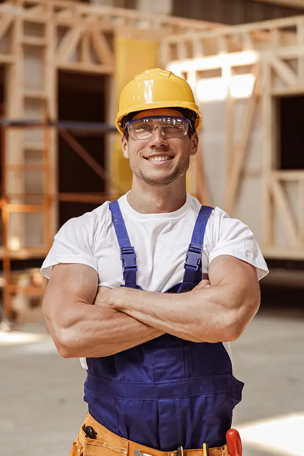 A construction worker with a yellow hard hat and safety glasses is smiling. He is wearing a white t-shirt and blue overalls, standing with arms crossed in a building under construction. Wooden framework and scaffolding are visible in the background. Dominion Roofing | BRAMPTON | BURLINGTON | ETOBICOKE | MARKHAM | MISSISSAUGA | NORTH YORK | OAKVILLE | RICHMOND HILL | Scarborough | TORONTO | VAUGHAN | dominionroofing.com | Phone Number: 416-789-0601