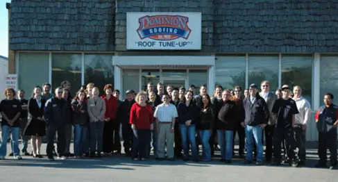 A large group of people stands in front of a building with a sign that reads Dominion Roofing Roof Tune-Up. They are smiling and posing for a group photo. Dominion Roofing | BRAMPTON | BURLINGTON | ETOBICOKE | MARKHAM | MISSISSAUGA | NORTH YORK | OAKVILLE | RICHMOND HILL | Scarborough | TORONTO | VAUGHAN | dominionroofing.com | Phone Number: 416-789-0601