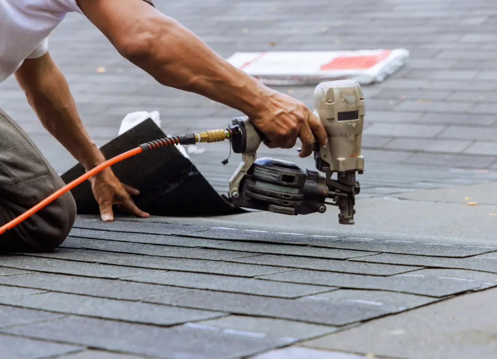 A person using a nail gun to install asphalt shingles on a roof, with a focus on the tool and shingles. The persons arm is visible, wearing a white shirt. Shingles are stacked in the background. Dominion Roofing | BRAMPTON | BURLINGTON | ETOBICOKE | MARKHAM | MISSISSAUGA | NORTH YORK | OAKVILLE | RICHMOND HILL | Scarborough | TORONTO | VAUGHAN | dominionroofing.com | Phone Number: 416-789-0601