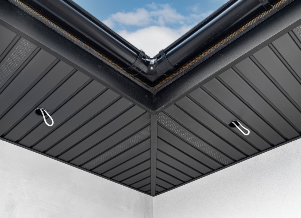 View of a modern roof corner with black metallic panels and two hanging loops. The roof features a downspout at the corner, and the sky is visible in the background. The design is clean and geometric. Dominion Roofing | BRAMPTON | BURLINGTON | ETOBICOKE | MARKHAM | MISSISSAUGA | NORTH YORK | OAKVILLE | RICHMOND HILL | Scarborough | TORONTO | VAUGHAN | dominionroofing.com | Phone Number: 416-789-0601