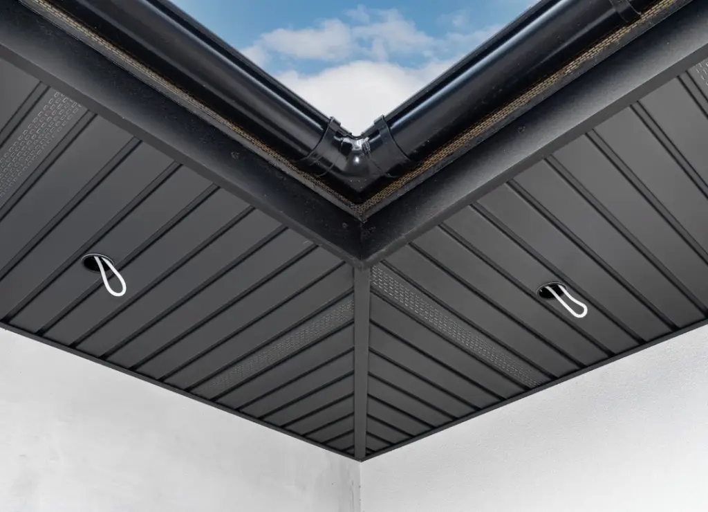 View of a modern roof corner with black metallic panels and two hanging loops. The roof features a downspout at the corner, and the sky is visible in the background. The design is clean and geometric. Dominion Roofing | BRAMPTON | BURLINGTON | ETOBICOKE | MARKHAM | MISSISSAUGA | NORTH YORK | OAKVILLE | RICHMOND HILL | Scarborough | TORONTO | VAUGHAN | dominionroofing.com | Phone Number: 416-789-0601