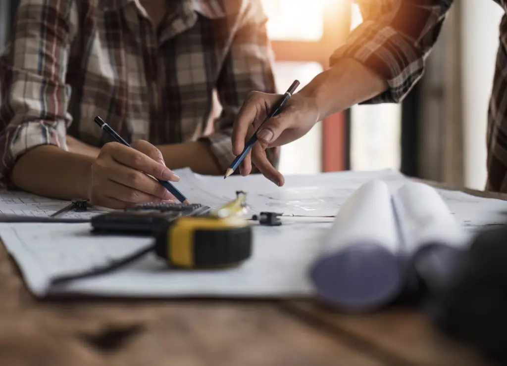 Two people collaborate over architectural blueprints on a wooden table. One person points with a pencil, while the other holds a pencil, wearing plaid shirts. A tape measure and rolled-up papers are also visible. Sunlight filters in from a window. Dominion Roofing | BRAMPTON | BURLINGTON | ETOBICOKE | MARKHAM | MISSISSAUGA | NORTH YORK | OAKVILLE | RICHMOND HILL | Scarborough | TORONTO | VAUGHAN | dominionroofing.com | Phone Number: 416-789-0601