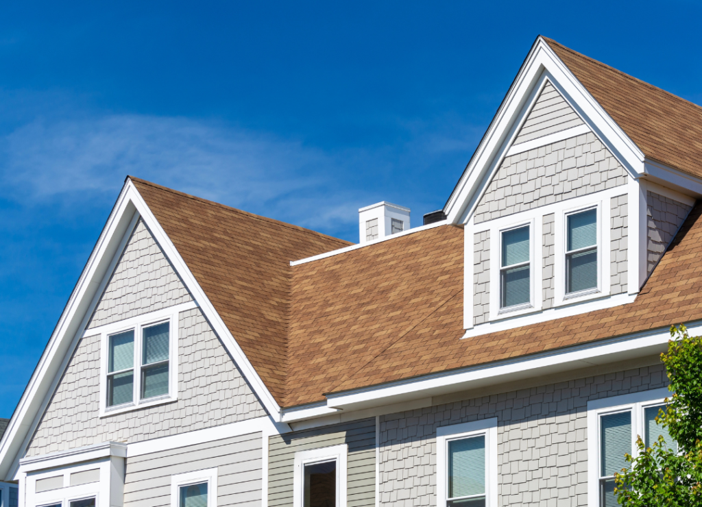 A beige and white two-story house with brown shingle roof against a clear blue sky. The exterior features decorative white trim, large windows with white frames, and tree foliage visible at the bottom right corner. Dominion Roofing | BRAMPTON | BURLINGTON | ETOBICOKE | MARKHAM | MISSISSAUGA | NORTH YORK | OAKVILLE | RICHMOND HILL | Scarborough | TORONTO | VAUGHAN | dominionroofing.com | Phone Number: 416-789-0601
