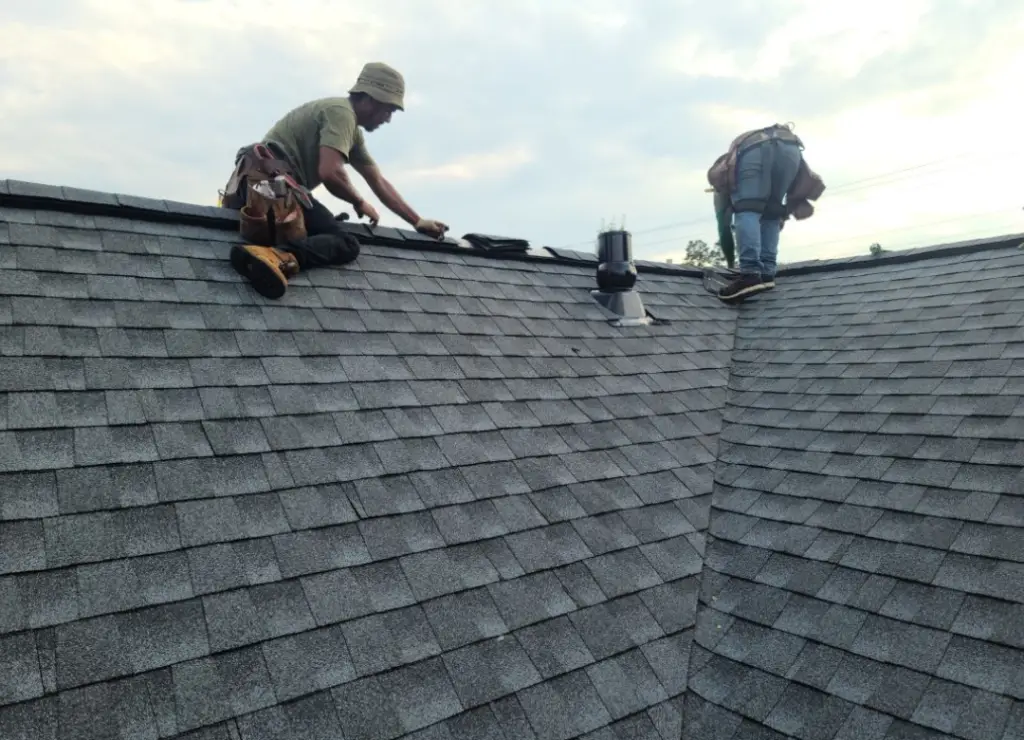 Two workers on a pitched gray shingle roof; one is kneeling, using a tool while the other is standing, carrying equipment. Both wear safety gear as they work under a cloudy sky. Dominion Roofing | BRAMPTON | BURLINGTON | ETOBICOKE | MARKHAM | MISSISSAUGA | NORTH YORK | OAKVILLE | RICHMOND HILL | Scarborough | TORONTO | VAUGHAN | dominionroofing.com | Phone Number: 416-789-0601
