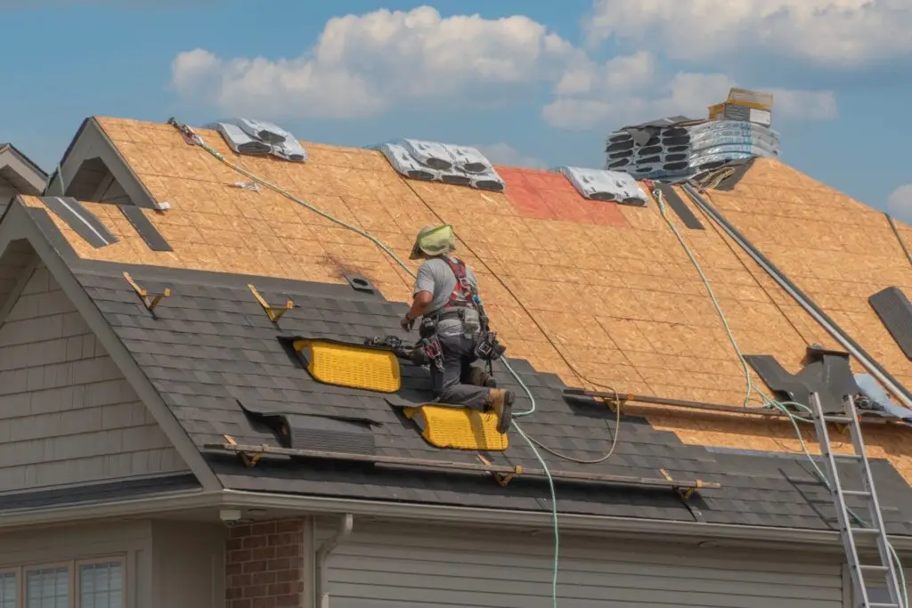 A construction worker is installing shingles on a house roof under a partly cloudy sky. They are wearing safety gear and using tools, with bundles of shingles and materials visible on the roof. Dominion Roofing | BRAMPTON | BURLINGTON | ETOBICOKE | MARKHAM | MISSISSAUGA | NORTH YORK | OAKVILLE | RICHMOND HILL | Scarborough | TORONTO | VAUGHAN | dominionroofing.com | Phone Number: 416-789-0601