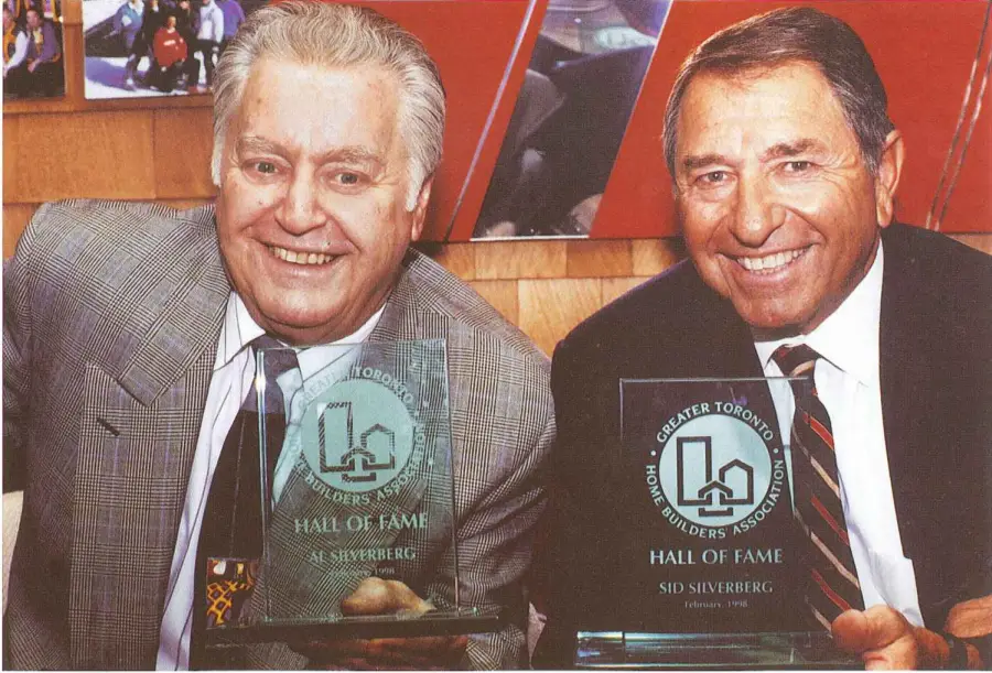 Two men smiling and holding crystal awards from the Greater Toronto Home Builders Association Hall of Fame. Both are wearing suits and ties, standing against a backdrop featuring a wood panel and abstract art. Dominion Roofing | BRAMPTON | BURLINGTON | ETOBICOKE | MARKHAM | MISSISSAUGA | NORTH YORK | OAKVILLE | RICHMOND HILL | Scarborough | TORONTO | VAUGHAN | dominionroofing.com | Phone Number: 416-789-0601