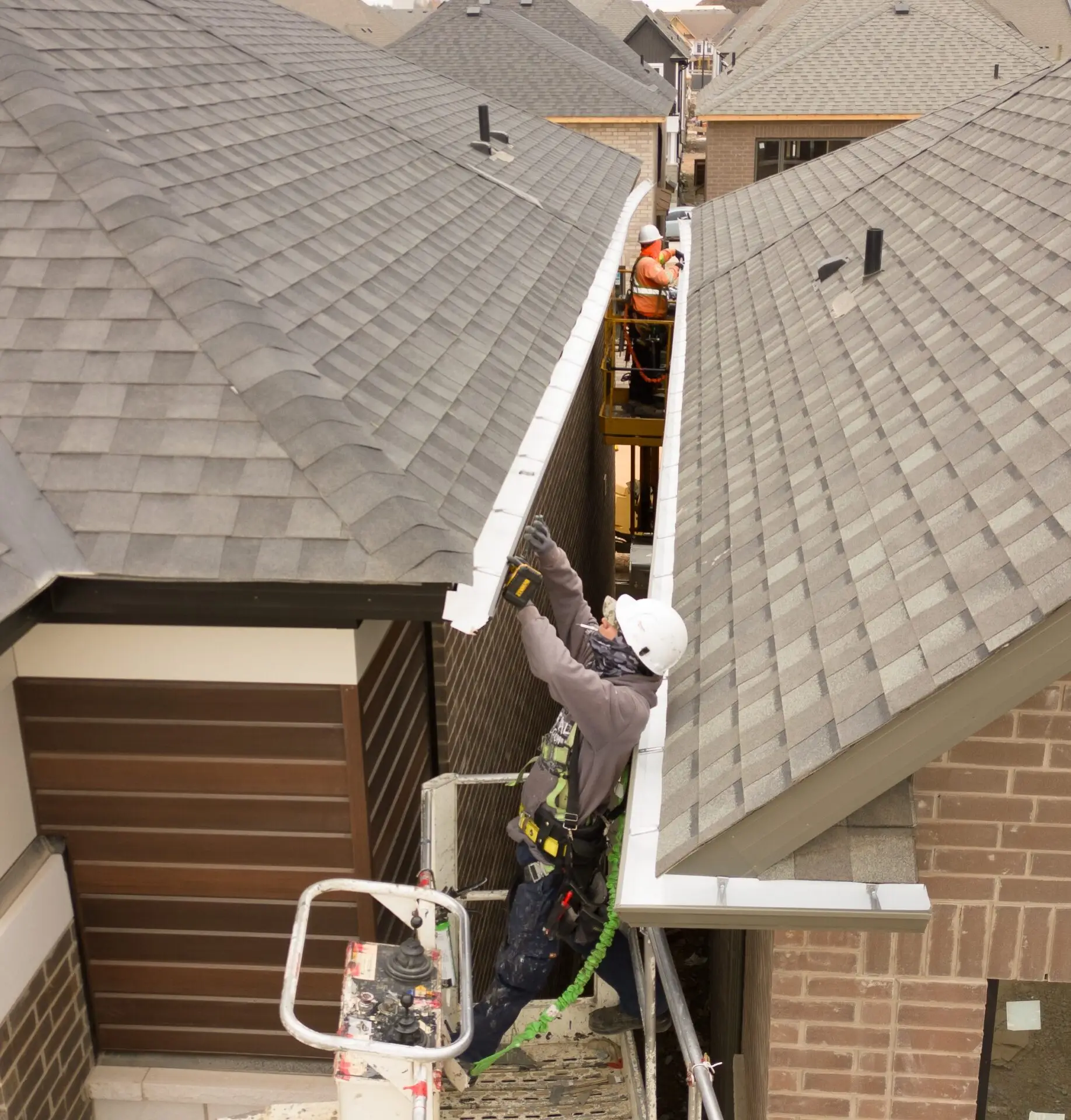 A worker in a hard hat and safety harness installs residential eavestroughs on a roof while standing on scaffolding. In the background, multiple houses with similar shingle roofs showcase the completed downpipe services. Dominion Roofing | BRAMPTON | BURLINGTON | ETOBICOKE | MARKHAM | MISSISSAUGA | NORTH YORK | OAKVILLE | RICHMOND HILL | Scarborough | TORONTO | VAUGHAN | dominionroofing.com | Phone Number: 416-789-0601