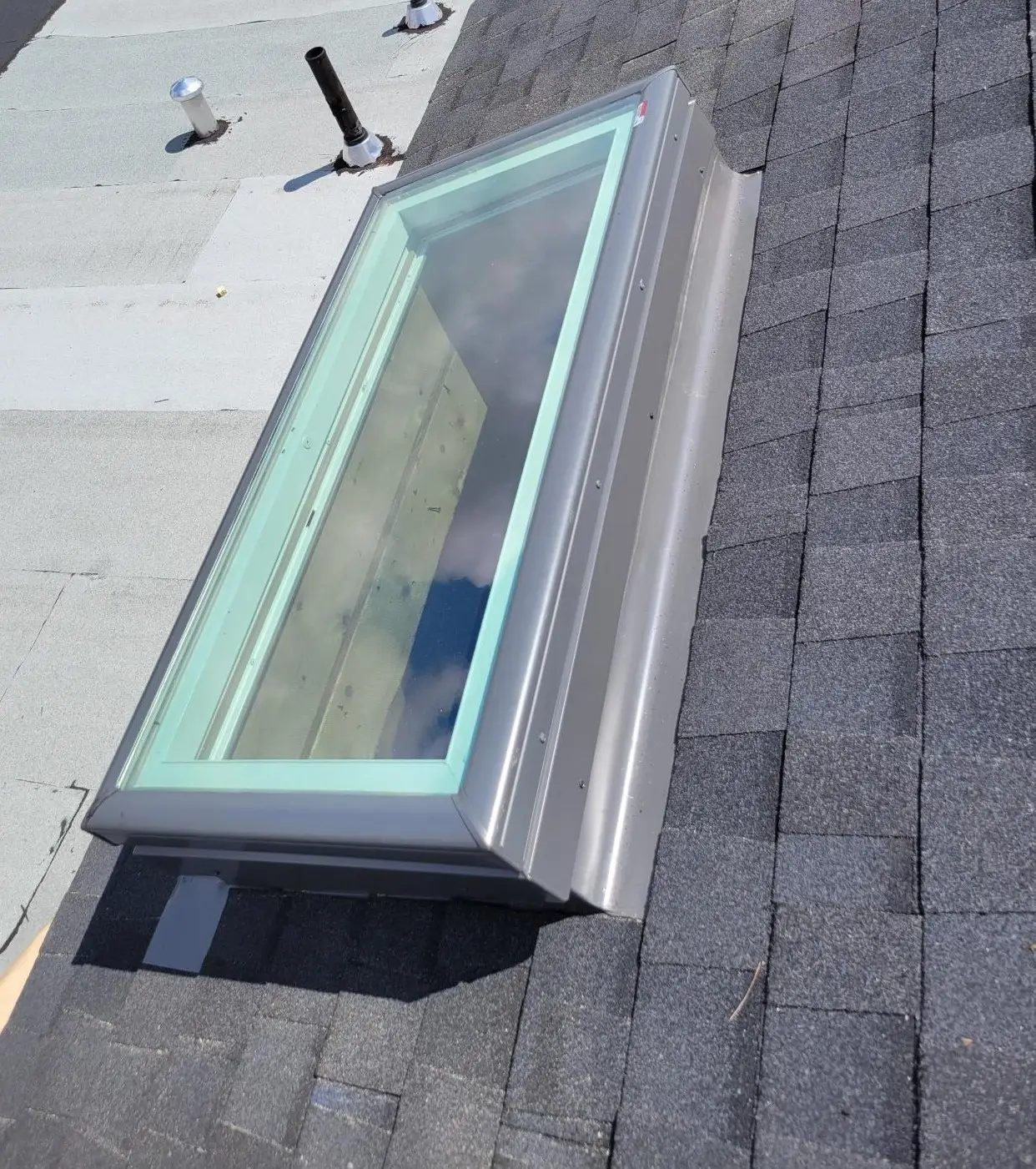 A skylight installed on a roof, partially surrounded by shingles. The glass is clear, revealing a reflection of the sky. The roof features a combination of darker shingles and lighter flat materials. Two pipe vents are visible nearby. Dominion Roofing | BRAMPTON | BURLINGTON | ETOBICOKE | MARKHAM | MISSISSAUGA | NORTH YORK | OAKVILLE | RICHMOND HILL | Scarborough | TORONTO | VAUGHAN | dominionroofing.com | Phone Number: 416-789-0601