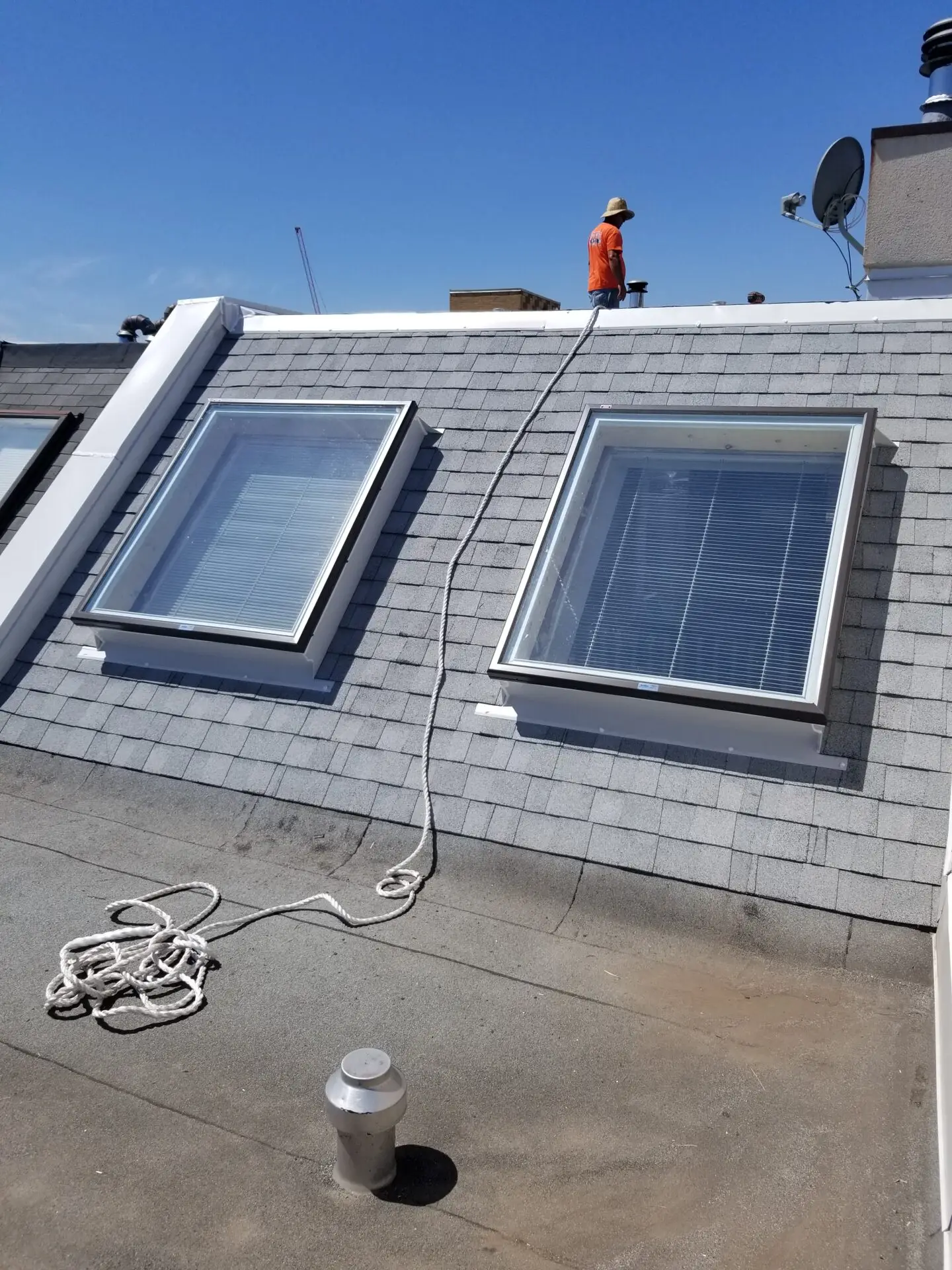 A worker in an orange shirt and helmet is standing on a rooftop with gray shingles. Two skylights with closed blinds are visible beside him. A rope is coiled on the flat part of the roof. The sky is clear and blue. Dominion Roofing | BRAMPTON | BURLINGTON | ETOBICOKE | MARKHAM | MISSISSAUGA | NORTH YORK | OAKVILLE | RICHMOND HILL | Scarborough | TORONTO | VAUGHAN | dominionroofing.com | Phone Number: 416-789-0601