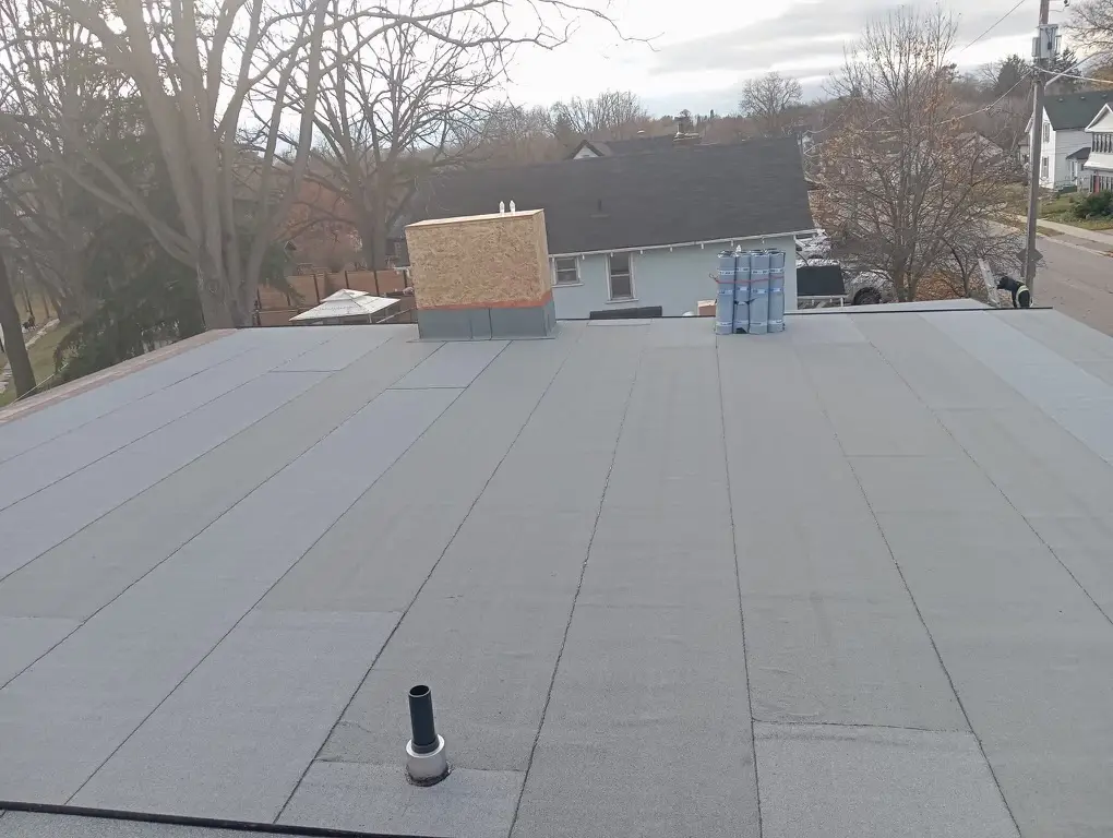 A flat gray rooftop, featuring a chimney covered in plywood and two vent pipes, suggests new construction roofing for homes. A stack of insulation sheets is visible, while leafless trees and residential houses stretch into the background under a cloudy sky. Dominion Roofing | BRAMPTON | BURLINGTON | ETOBICOKE | MARKHAM | MISSISSAUGA | NORTH YORK | OAKVILLE | RICHMOND HILL | Scarborough | TORONTO | VAUGHAN | dominionroofing.com | Phone Number: 416-789-0601