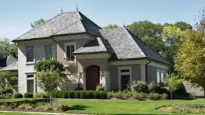 A modern two-story house with a sloped gray slate roof, beige walls, and a prominent red front door. Surrounding the house are well-manicured shrubs and small trees, set against a backdrop of lush greenery and trees. Dominion Roofing | BRAMPTON | BURLINGTON | ETOBICOKE | MARKHAM | MISSISSAUGA | NORTH YORK | OAKVILLE | RICHMOND HILL | Scarborough | TORONTO | VAUGHAN | dominionroofing.com | Phone Number: 416-789-0601
