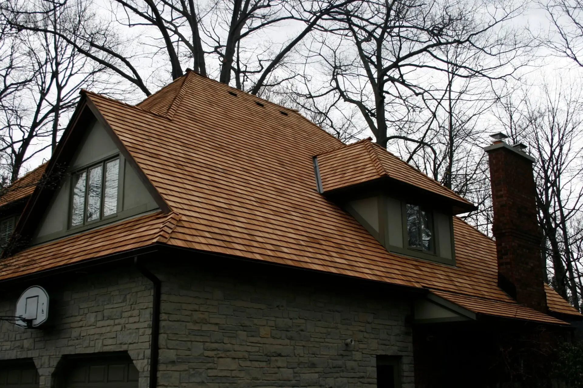 A house with a steep, brown wooden shingle roof, stone exterior walls, and large windows is surrounded by tall, bare trees. A basketball hoop is attached to the garage in the foreground. The sky is overcast. Dominion Roofing | BRAMPTON | BURLINGTON | ETOBICOKE | MARKHAM | MISSISSAUGA | NORTH YORK | OAKVILLE | RICHMOND HILL | Scarborough | TORONTO | VAUGHAN | dominionroofing.com | Phone Number: 416-789-0601