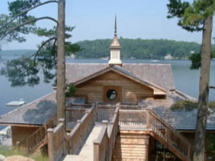 A rustic wooden building with residential shingle roofing and a pointed spire, surrounded by tall trees, overlooks a serene lake. A wooden walkway with railings leads to the structure. Forested hills are visible in the background under a clear sky. Dominion Roofing | BRAMPTON | BURLINGTON | ETOBICOKE | MARKHAM | MISSISSAUGA | NORTH YORK | OAKVILLE | RICHMOND HILL | Scarborough | TORONTO | VAUGHAN | dominionroofing.com | Phone Number: 416-789-0601
