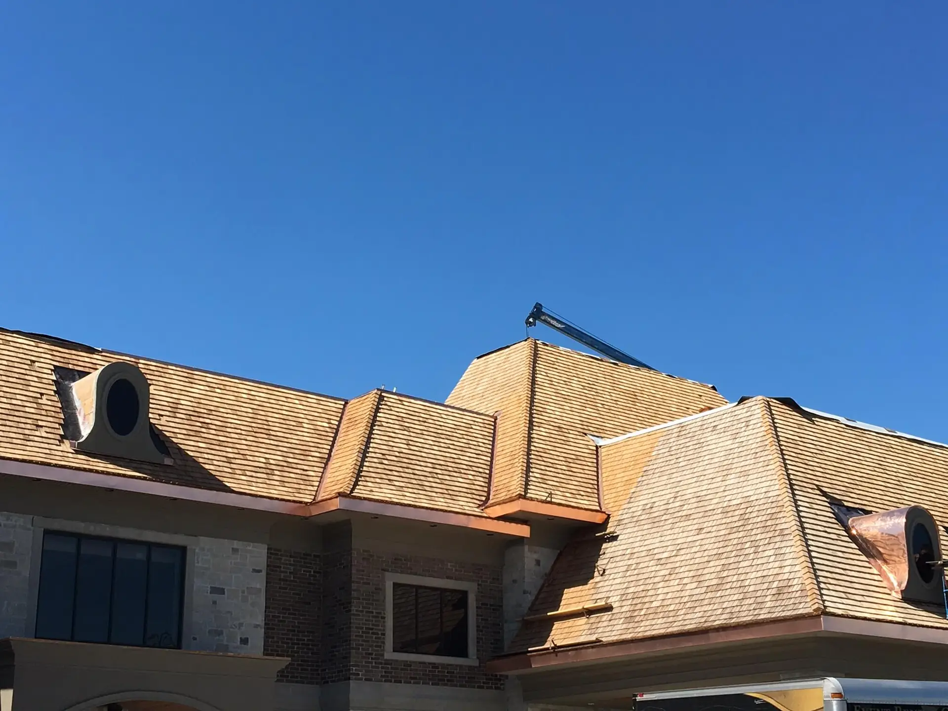 A building with a complex light brown shingle roof features multiple gables and circular dormer windows against a clear blue sky. Dominion Roofing | BRAMPTON | BURLINGTON | ETOBICOKE | MARKHAM | MISSISSAUGA | NORTH YORK | OAKVILLE | RICHMOND HILL | Scarborough | TORONTO | VAUGHAN | dominionroofing.com | Phone Number: 416-789-0601
