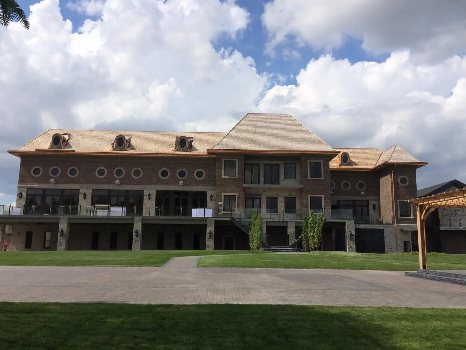 A large, elegant brick building with multiple windows and dormers under a cloudy blue sky, showcasing its specialty commercial roofing solutions. The structure is surrounded by neatly manicured grass and a paved area in the foreground. Dominion Roofing | BRAMPTON | BURLINGTON | ETOBICOKE | MARKHAM | MISSISSAUGA | NORTH YORK | OAKVILLE | RICHMOND HILL | Scarborough | TORONTO | VAUGHAN | dominionroofing.com | Phone Number: 416-789-0601