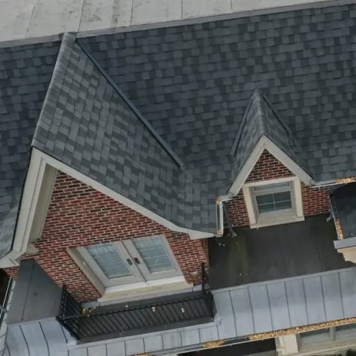 Brick building with a black shingled roof recently updated through residential roof replacement. A small balcony features two sets of glass doors and a black metal railing. The roofs varying angles and shades create a dynamic, multi-level appearance. Dominion Roofing | BRAMPTON | BURLINGTON | ETOBICOKE | MARKHAM | MISSISSAUGA | NORTH YORK | OAKVILLE | RICHMOND HILL | Scarborough | TORONTO | VAUGHAN | dominionroofing.com | Phone Number: 416-789-0601