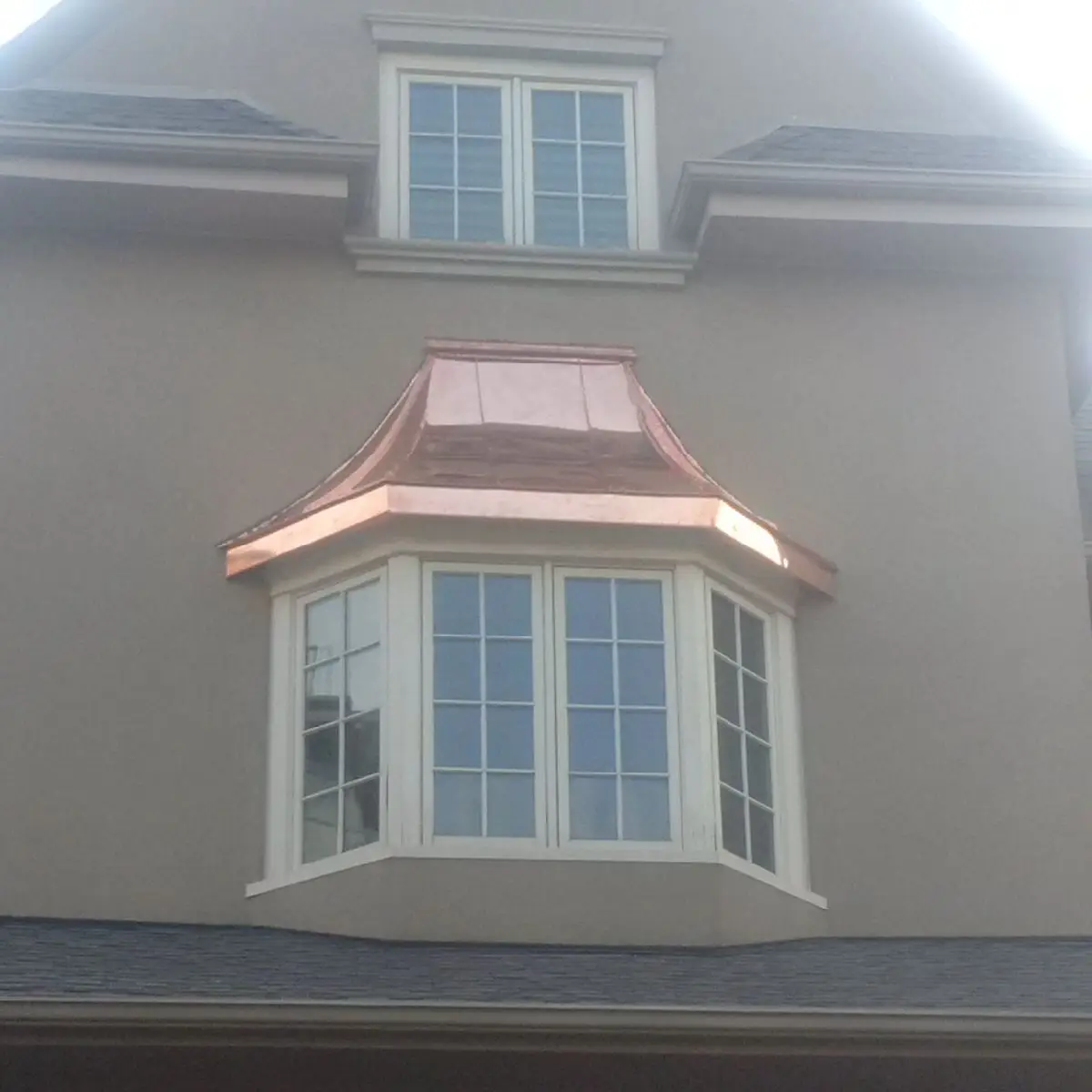A two-story house features a window with four panels on the second floor, topped with a new, shiny copper roof. Above, there is a smaller, square window with four sections. The house exterior is a light beige color. Dominion Roofing | BRAMPTON | BURLINGTON | ETOBICOKE | MARKHAM | MISSISSAUGA | NORTH YORK | OAKVILLE | RICHMOND HILL | Scarborough | TORONTO | VAUGHAN | dominionroofing.com | Phone Number: 416-789-0601