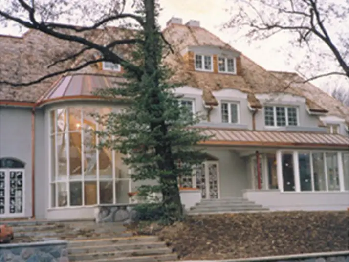 A large, elegant house with residential shingle roofing features numerous windows and a glass-walled section. Surrounded by bare trees, stairs leading up to it, and a stone base, the scene evokes a classic, cozy architectural style. Dominion Roofing | BRAMPTON | BURLINGTON | ETOBICOKE | MARKHAM | MISSISSAUGA | NORTH YORK | OAKVILLE | RICHMOND HILL | Scarborough | TORONTO | VAUGHAN | dominionroofing.com | Phone Number: 416-789-0601