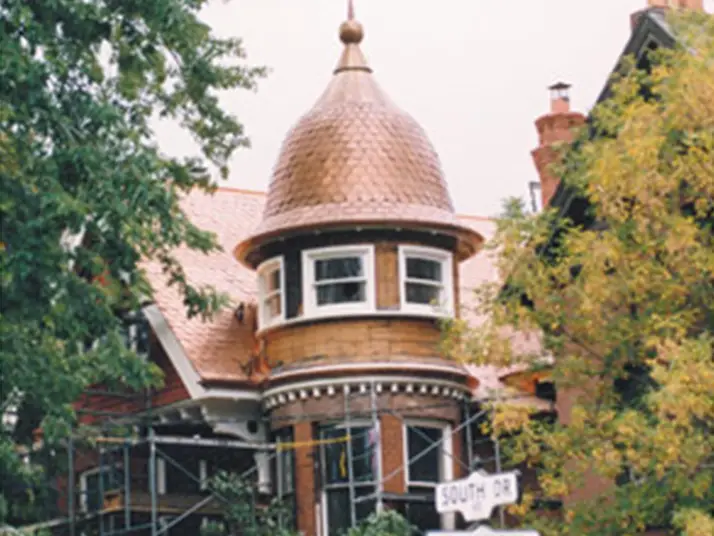 A Victorian-style house with a conical turret roof and large windows, partially obscured by trees, features residential shingle roofing in progress. Scaffolding surrounds the renovation site. In the foreground, a street sign reads South Dr. Dominion Roofing | BRAMPTON | BURLINGTON | ETOBICOKE | MARKHAM | MISSISSAUGA | NORTH YORK | OAKVILLE | RICHMOND HILL | Scarborough | TORONTO | VAUGHAN | dominionroofing.com | Phone Number: 416-789-0601