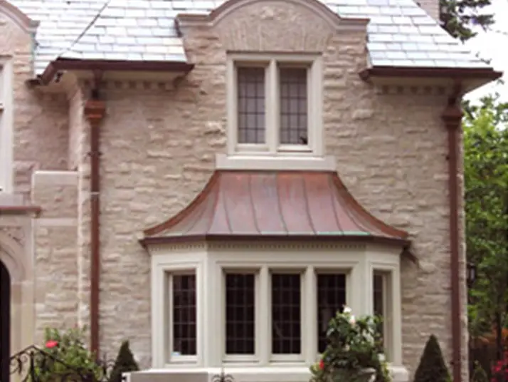 A stone house with a bay window and copper roof is surrounded by greenery. The upper windows boast decorative panes, while the blue-gray residential shingle roofing adds charm. The exterior wall showcases textured stonework. Dominion Roofing | BRAMPTON | BURLINGTON | ETOBICOKE | MARKHAM | MISSISSAUGA | NORTH YORK | OAKVILLE | RICHMOND HILL | Scarborough | TORONTO | VAUGHAN | dominionroofing.com | Phone Number: 416-789-0601
