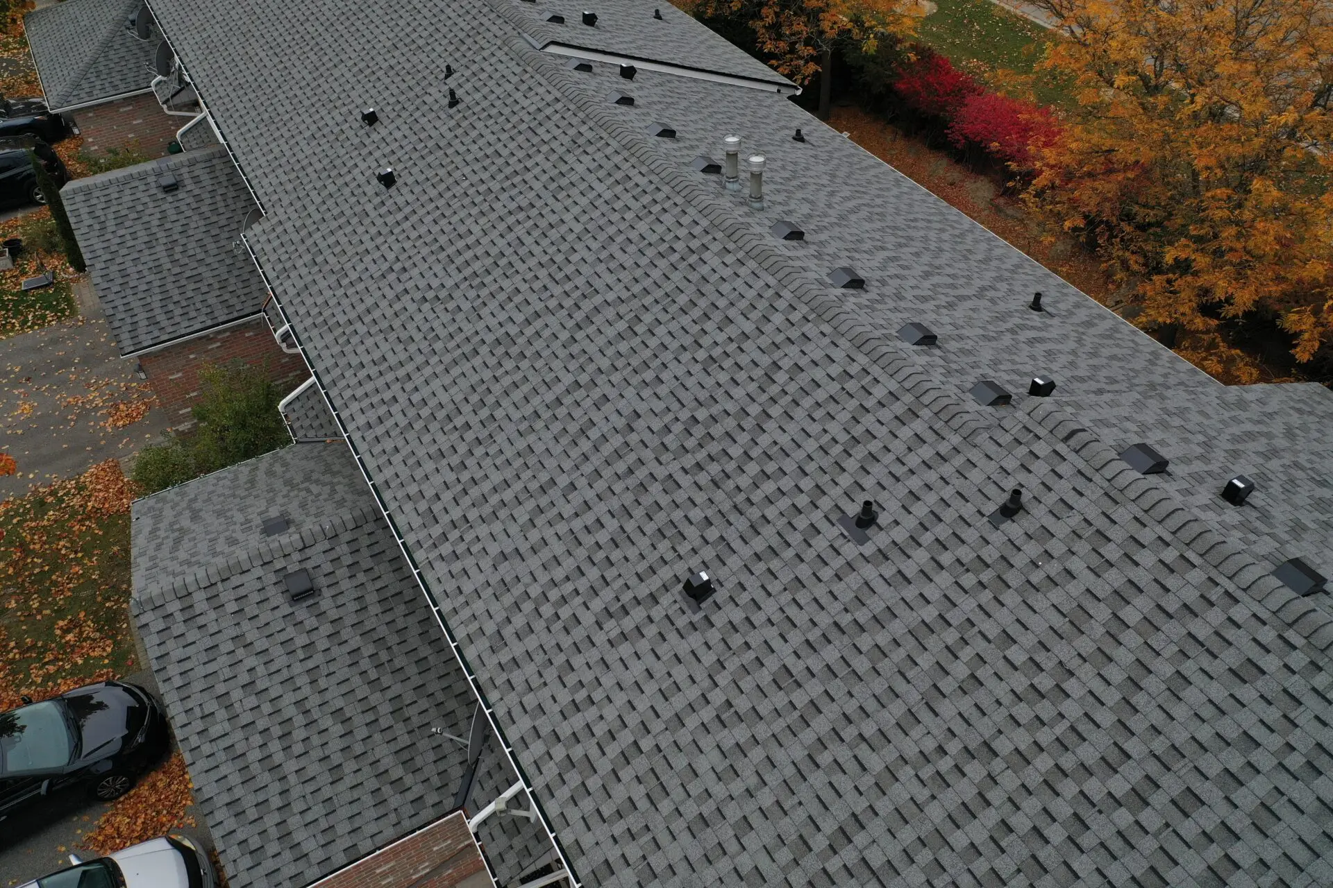 Aerial view of a large, gray shingled roof on a residential building, showcasing the quality of our commercial shingle roofing services. Surrounded by autumn trees with orange and red leaves, several cars are parked nearby. Vent pipes and chimney stacks are visible on the roof. Dominion Roofing | BRAMPTON | BURLINGTON | ETOBICOKE | MARKHAM | MISSISSAUGA | NORTH YORK | OAKVILLE | RICHMOND HILL | Scarborough | TORONTO | VAUGHAN | dominionroofing.com | Phone Number: 416-789-0601