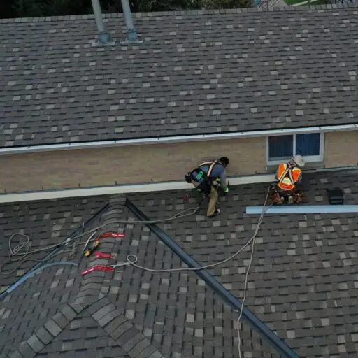 Two workers wearing safety gear and helmets are undertaking a residential roof replacement on a sloped shingle roof. They are installing or repairing equipment near the roofs edge, with multiple ropes and tools visible, including a large pole securely attached to the structure. Dominion Roofing | BRAMPTON | BURLINGTON | ETOBICOKE | MARKHAM | MISSISSAUGA | NORTH YORK | OAKVILLE | RICHMOND HILL | Scarborough | TORONTO | VAUGHAN | dominionroofing.com | Phone Number: 416-789-0601