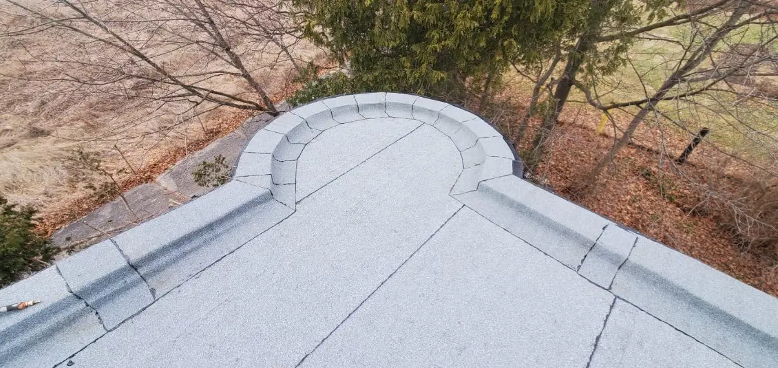 A rooftop view showcases a semicircular stone parapet at the edge, typical of residential flat roofing, surrounded by bare trees and fallen leaves. The scene overlooks a blend of grass and leaf-covered ground in the outdoor area below. Dominion Roofing | BRAMPTON | BURLINGTON | ETOBICOKE | MARKHAM | MISSISSAUGA | NORTH YORK | OAKVILLE | RICHMOND HILL | Scarborough | TORONTO | VAUGHAN | dominionroofing.com | Phone Number: 416-789-0601