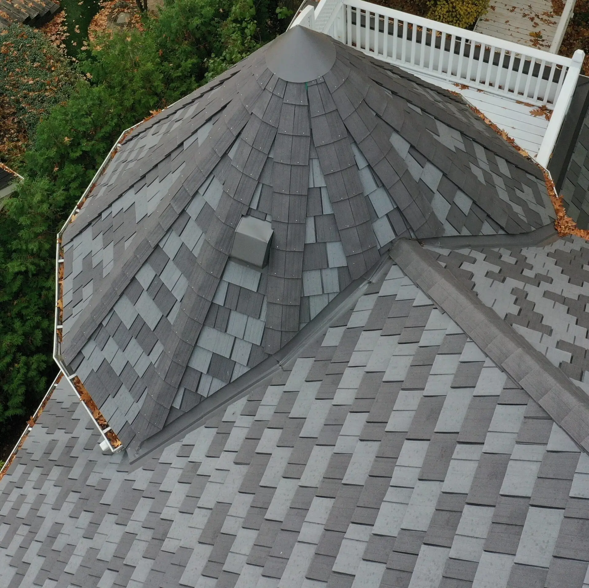 Aerial view of a gray shingled roof, showcasing specialty residential roofing with its multi-sided, conical shape and vent. Surrounded by lush greenery, a white balcony railing peeks out at the top right corner. Dominion Roofing | BRAMPTON | BURLINGTON | ETOBICOKE | MARKHAM | MISSISSAUGA | NORTH YORK | OAKVILLE | RICHMOND HILL | Scarborough | TORONTO | VAUGHAN | dominionroofing.com | Phone Number: 416-789-0601