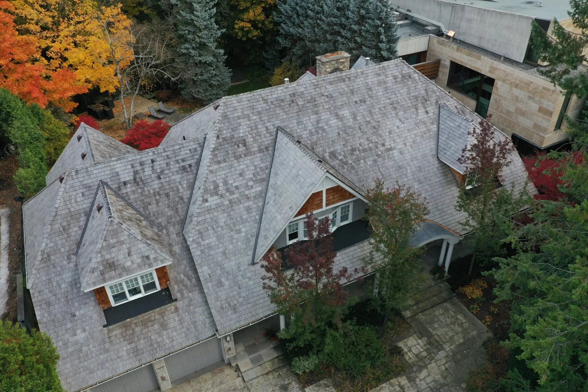 An aerial view of a large house showcases its specialty residential roofing with a gray, intricate design. Surrounded by the vibrant hues of autumn trees, the front features a welcoming porch and multiple windows, set amidst red and orange foliage that beautifully frames the property. Dominion Roofing | BRAMPTON | BURLINGTON | ETOBICOKE | MARKHAM | MISSISSAUGA | NORTH YORK | OAKVILLE | RICHMOND HILL | Scarborough | TORONTO | VAUGHAN | dominionroofing.com | Phone Number: 416-789-0601