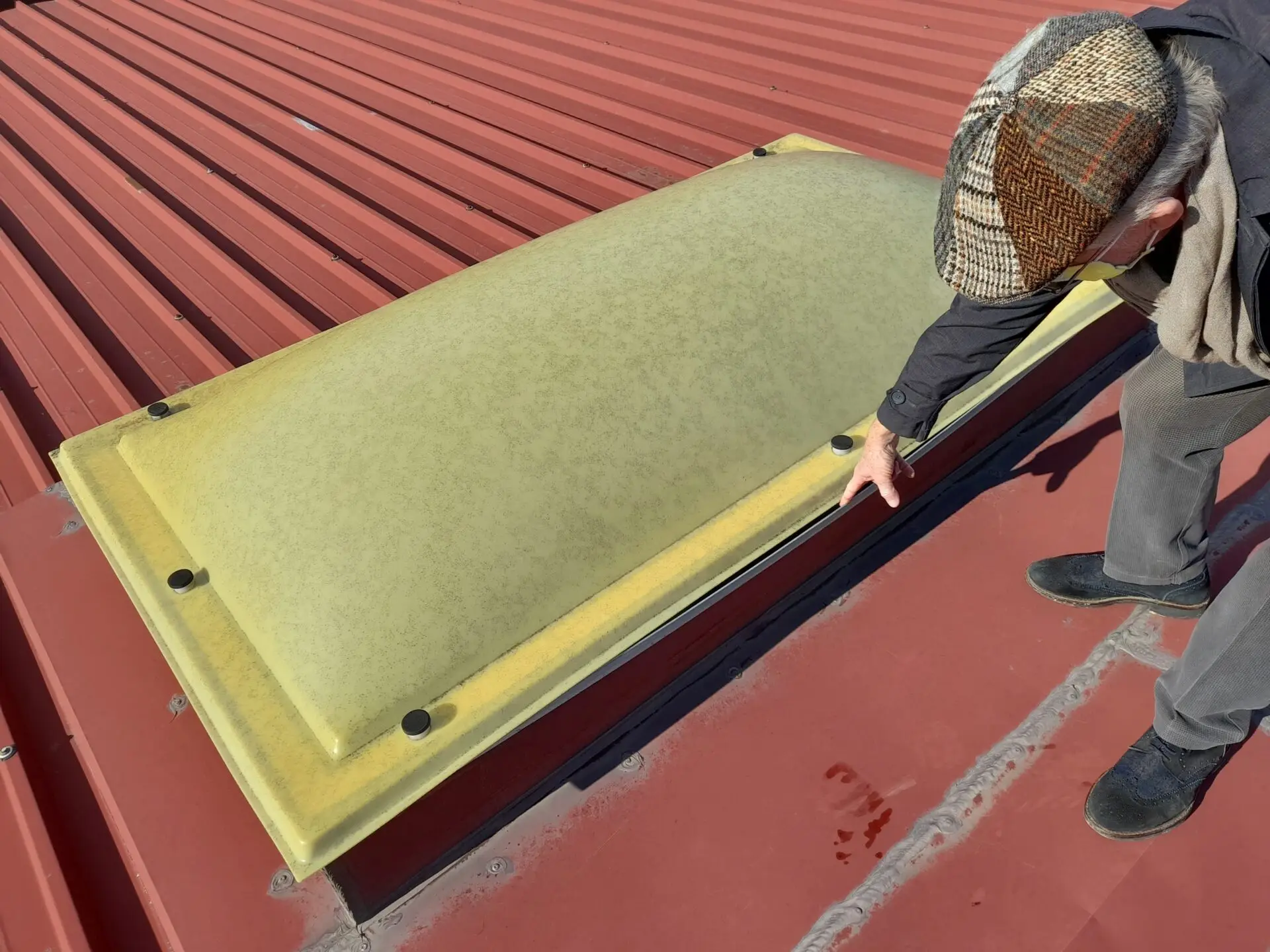 Person pointing at a large, rectangular, yellow-tinted skylight on a red metal roof. The skylight is fastened with black bolts. The person is wearing a checkered hat and a gray jacket, and the sun casts a shadow on the roof. Dominion Roofing | BRAMPTON | BURLINGTON | ETOBICOKE | MARKHAM | MISSISSAUGA | NORTH YORK | OAKVILLE | RICHMOND HILL | Scarborough | TORONTO | VAUGHAN | dominionroofing.com | Phone Number: 416-789-0601