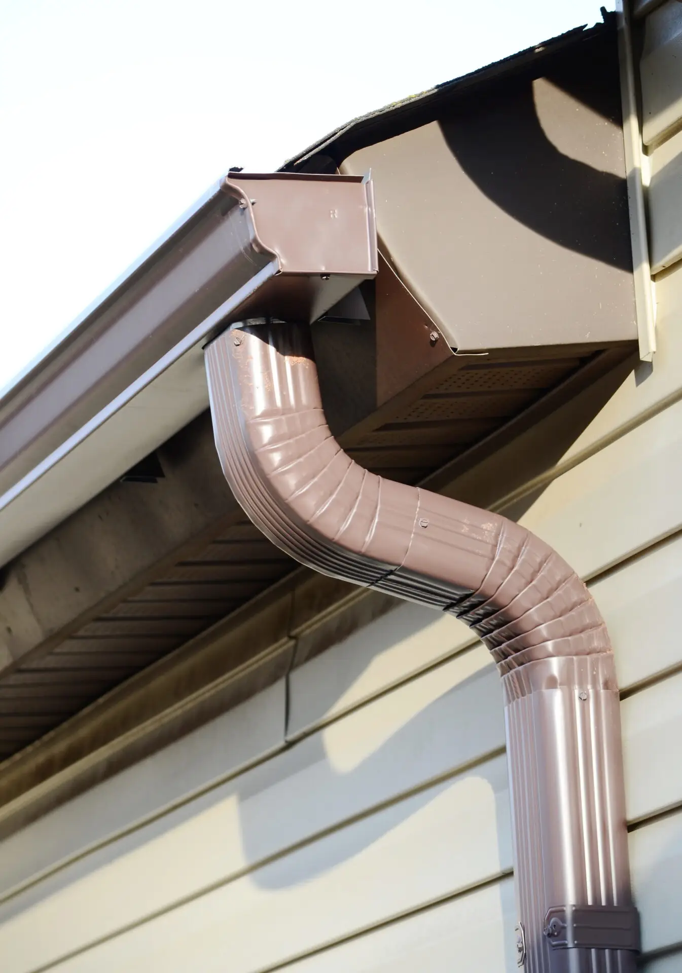 A close-up of a brown rain gutter and downspout, part of a residential eavestrough system, attached to the corner of a house with siding. Sunlight casts shadows, highlighting the metallic texture and the elegant curve of the downpipe. Dominion Roofing | BRAMPTON | BURLINGTON | ETOBICOKE | MARKHAM | MISSISSAUGA | NORTH YORK | OAKVILLE | RICHMOND HILL | Scarborough | TORONTO | VAUGHAN | dominionroofing.com | Phone Number: 416-789-0601