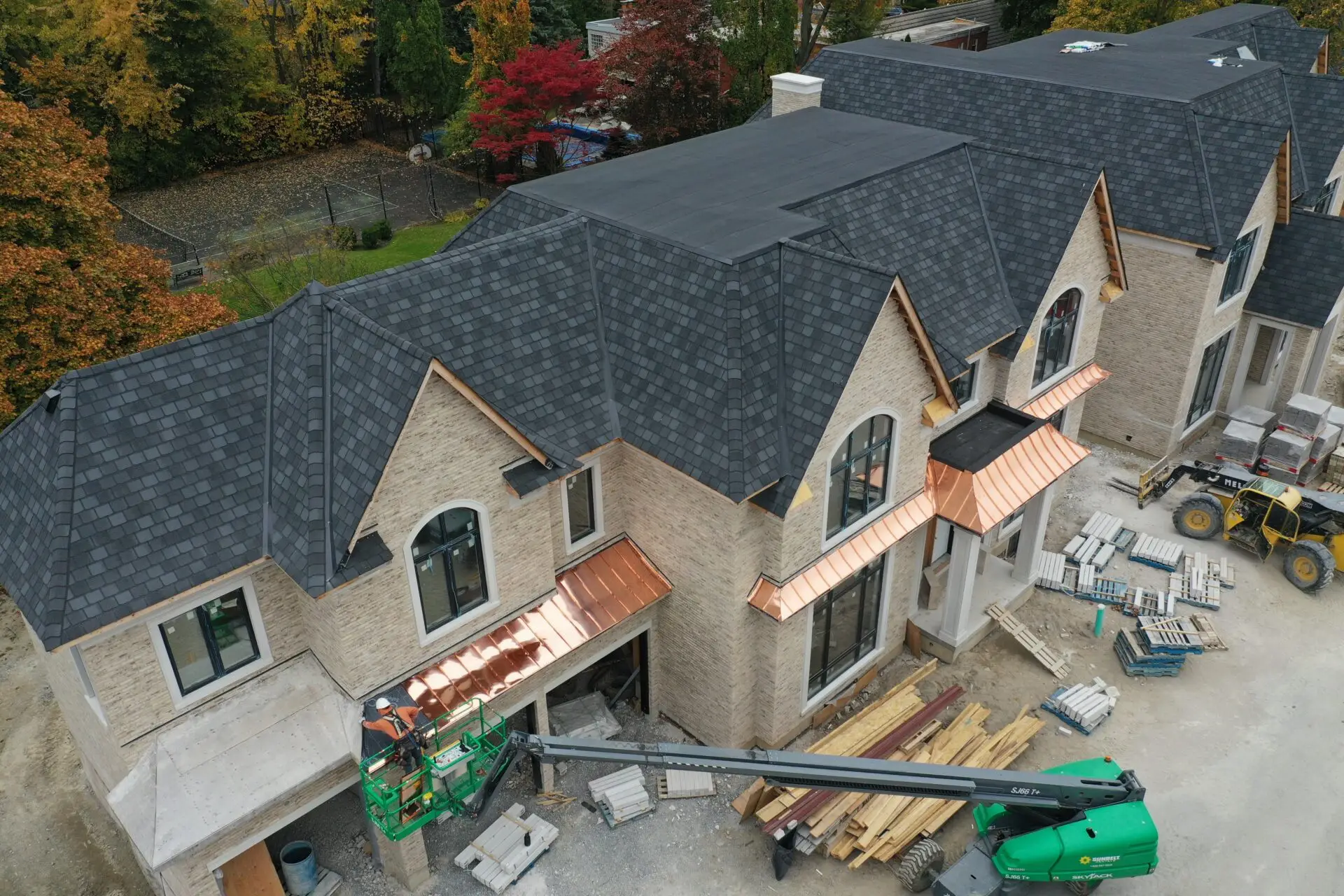 Aerial view of two large houses with grey shingle roofs, showcasing new construction roofing for commercial properties. Copper awnings glint in the sunlight. A green crane stands at the entrance amidst building materials and machinery, while trees with fall foliage provide a vibrant backdrop. Dominion Roofing | BRAMPTON | BURLINGTON | ETOBICOKE | MARKHAM | MISSISSAUGA | NORTH YORK | OAKVILLE | RICHMOND HILL | Scarborough | TORONTO | VAUGHAN | dominionroofing.com | Phone Number: 416-789-0601