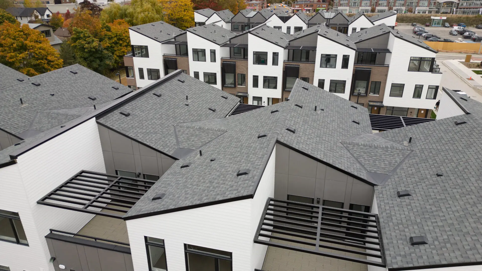 Aerial view of modern townhouses showcasing the sleek precision of new construction roofing, featuring angular gray roofs and white siding. The buildings boast large windows and wooden pergolas over the entrances, with autumn foliage and neighboring homes in the background. Dominion Roofing | BRAMPTON | BURLINGTON | ETOBICOKE | MARKHAM | MISSISSAUGA | NORTH YORK | OAKVILLE | RICHMOND HILL | Scarborough | TORONTO | VAUGHAN | dominionroofing.com | Phone Number: 416-789-0601