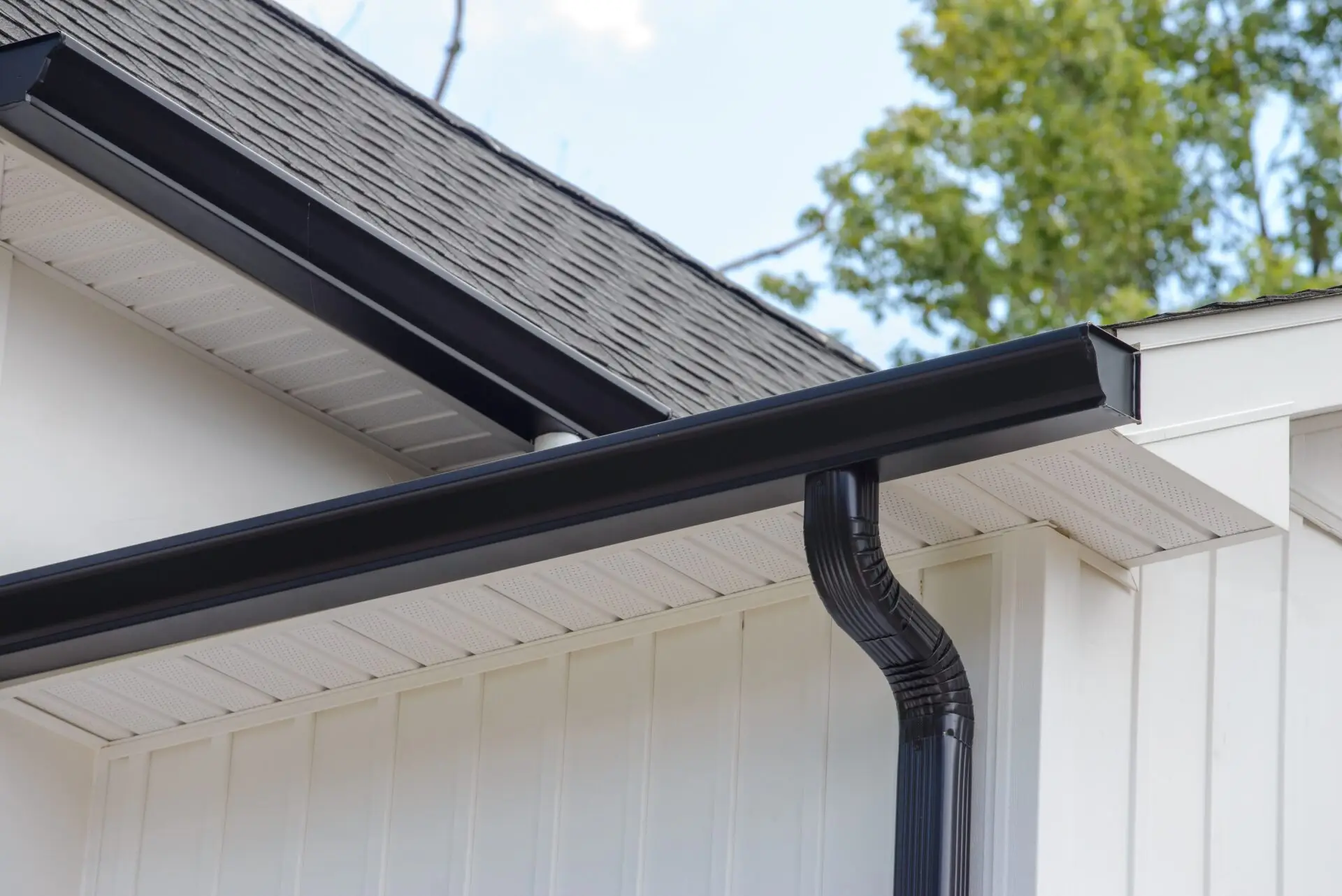 Residential eavestrough and downpipe services feature sleek black rain gutters installed on the edge of a buildings sloped, shingled roof. The gutters connect seamlessly to a downspout, running vertically along the white wooden siding. Trees and a blue sky provide a serene backdrop. Dominion Roofing | BRAMPTON | BURLINGTON | ETOBICOKE | MARKHAM | MISSISSAUGA | NORTH YORK | OAKVILLE | RICHMOND HILL | Scarborough | TORONTO | VAUGHAN | dominionroofing.com | Phone Number: 416-789-0601