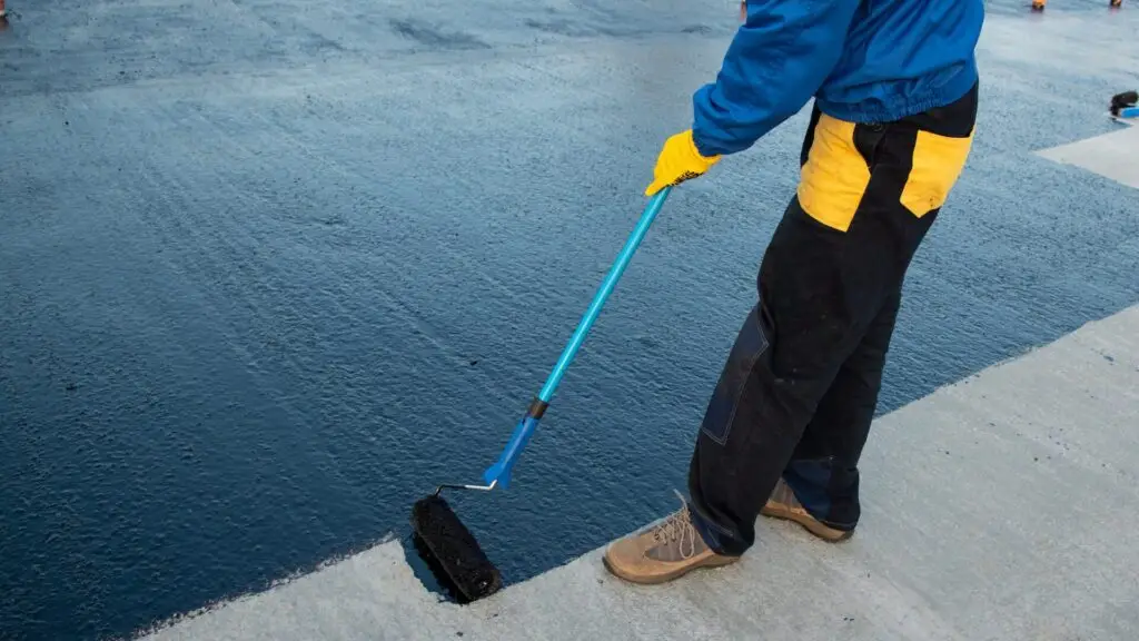 A person wearing a blue jacket, yellow gloves, and dark pants uses a long-handled roller to apply black sealant to a concrete surface. The ground appears partially covered, showing a contrast between sealed and unsealed areas. Dominion Roofing | BRAMPTON | BURLINGTON | ETOBICOKE | MARKHAM | MISSISSAUGA | NORTH YORK | OAKVILLE | RICHMOND HILL | Scarborough | TORONTO | VAUGHAN | dominionroofing.com | Phone Number: 416-789-0601