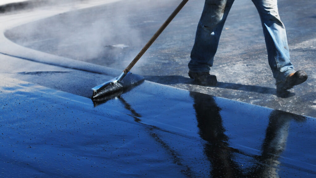 A person in jeans uses a long-handled tool to spread black tar for sealing or paving a surface. The tar is shiny and smooth, reflecting light, while steam or smoke rises from the hot material. Dominion Roofing | BRAMPTON | BURLINGTON | ETOBICOKE | MARKHAM | MISSISSAUGA | NORTH YORK | OAKVILLE | RICHMOND HILL | Scarborough | TORONTO | VAUGHAN | dominionroofing.com | Phone Number: 416-789-0601