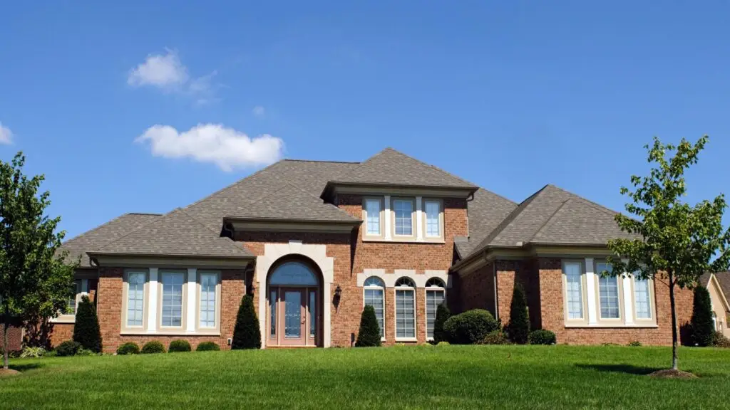 A large, two-story brick house with multiple windows and a double front door. The home has a gray roof and is surrounded by a well-maintained lawn and a few small trees under a clear blue sky. Dominion Roofing | BRAMPTON | BURLINGTON | ETOBICOKE | MARKHAM | MISSISSAUGA | NORTH YORK | OAKVILLE | RICHMOND HILL | Scarborough | TORONTO | VAUGHAN | dominionroofing.com | Phone Number: 416-789-0601