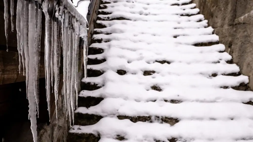 Snow-covered stone steps lead upward, with long icicles hanging from the railing on the left. The scene is wintry and cold, emphasizing the frozen environment. Dominion Roofing | BRAMPTON | BURLINGTON | ETOBICOKE | MARKHAM | MISSISSAUGA | NORTH YORK | OAKVILLE | RICHMOND HILL | Scarborough | TORONTO | VAUGHAN | dominionroofing.com | Phone Number: 416-789-0601