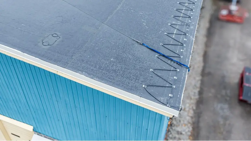 View of a buildings flat roof with safety cables and hooks along the edge. The building has blue walls and a white gutter. The ground below is blurred, with visible stones and a red object. Dominion Roofing | BRAMPTON | BURLINGTON | ETOBICOKE | MARKHAM | MISSISSAUGA | NORTH YORK | OAKVILLE | RICHMOND HILL | Scarborough | TORONTO | VAUGHAN | dominionroofing.com | Phone Number: 416-789-0601