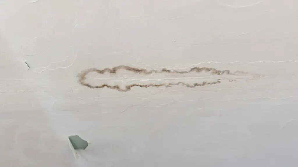 A damaged ceiling with a long, dark brown stain spreading outward, indicating a possible water leak. Near the stain, part of the ceilings surface is chipped away, revealing a hole. The ceiling is painted white with some cracks. Dominion Roofing | BRAMPTON | BURLINGTON | ETOBICOKE | MARKHAM | MISSISSAUGA | NORTH YORK | OAKVILLE | RICHMOND HILL | Scarborough | TORONTO | VAUGHAN | dominionroofing.com | Phone Number: 416-789-0601