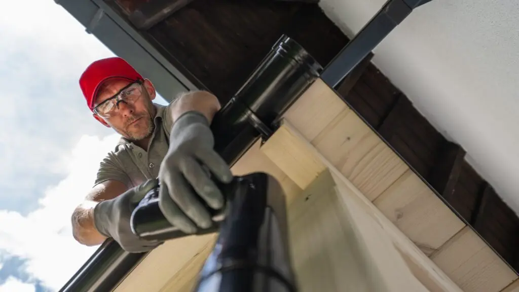 A person wearing a red cap, glasses, and gloves is installing or repairing a black gutter on a house. The photo is taken from below, looking up at the individual focused on their work. The sky is partly cloudy. Dominion Roofing | BRAMPTON | BURLINGTON | ETOBICOKE | MARKHAM | MISSISSAUGA | NORTH YORK | OAKVILLE | RICHMOND HILL | Scarborough | TORONTO | VAUGHAN | dominionroofing.com | Phone Number: 416-789-0601