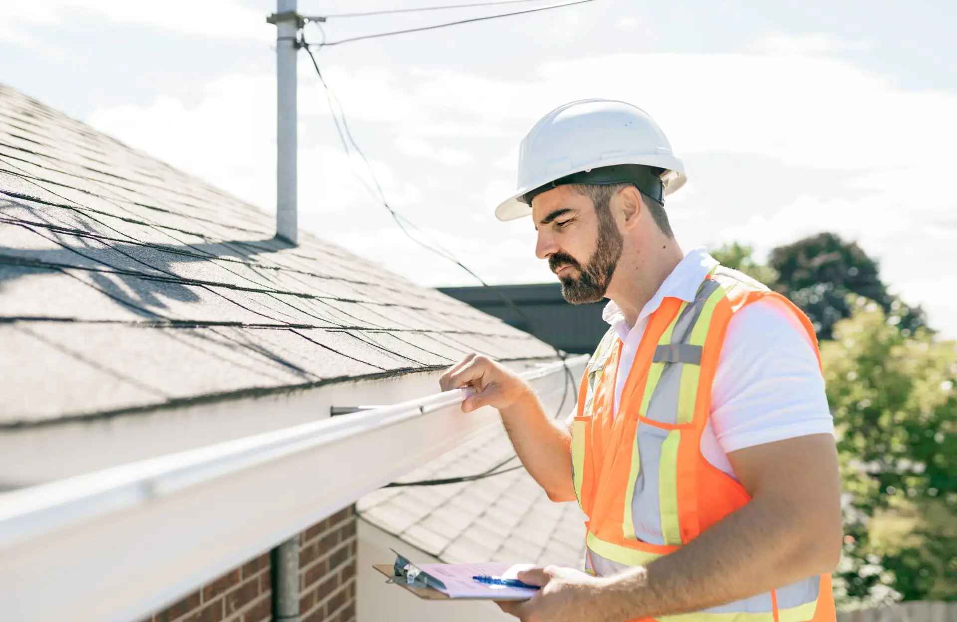 A construction worker wearing a white hard hat and orange safety vest inspects a gutter on the side of a house. He holds a clipboard and pen, standing under a clear blue sky. Dominion Roofing | BRAMPTON | BURLINGTON | ETOBICOKE | MARKHAM | MISSISSAUGA | NORTH YORK | OAKVILLE | RICHMOND HILL | Scarborough | TORONTO | VAUGHAN | dominionroofing.com | Phone Number: 416-789-0601