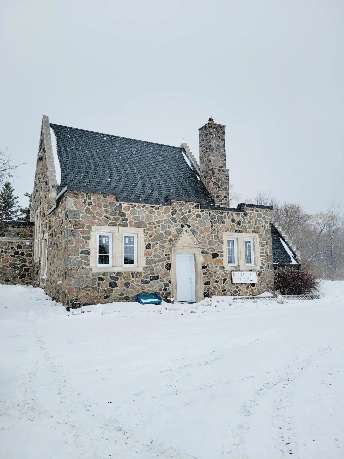 A stone house with a steep, dark roof crafted by Specialty Commercial Roofing Solutions sits in a snowy landscape. Snow covers the ground and bushes nearby, with tire tracks leading to the house. Bare trees are visible in the background under a gray sky. Dominion Roofing | BRAMPTON | BURLINGTON | ETOBICOKE | MARKHAM | MISSISSAUGA | NORTH YORK | OAKVILLE | RICHMOND HILL | Scarborough | TORONTO | VAUGHAN | dominionroofing.com | Phone Number: 416-789-0601