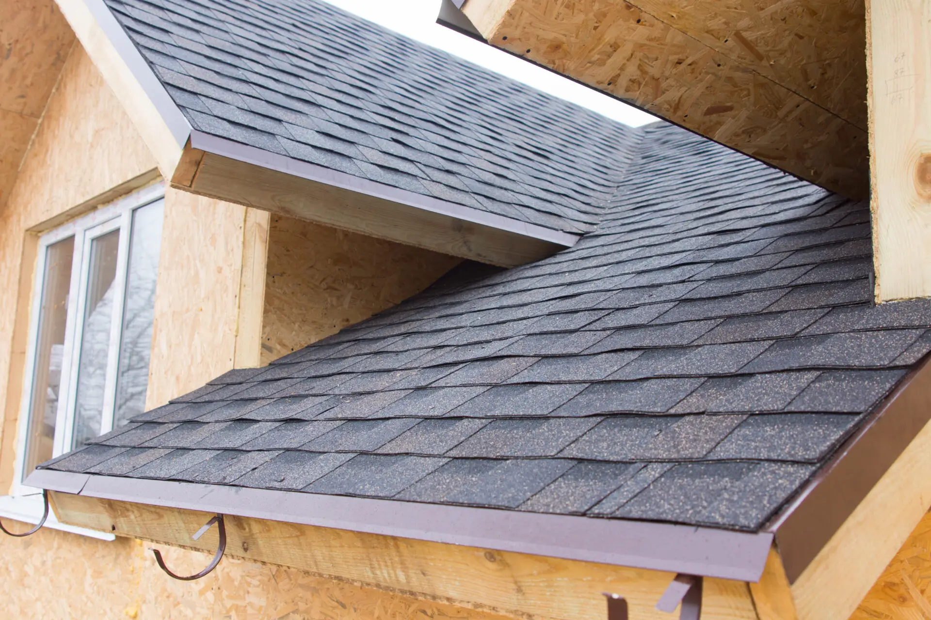 Close-up of a house with a dark shingled roof, showcasing the precision of new construction roofing. The roof features intersecting angles against plywood walls, while a window is partially visible on the left side. Dominion Roofing | BRAMPTON | BURLINGTON | ETOBICOKE | MARKHAM | MISSISSAUGA | NORTH YORK | OAKVILLE | RICHMOND HILL | Scarborough | TORONTO | VAUGHAN | dominionroofing.com | Phone Number: 416-789-0601