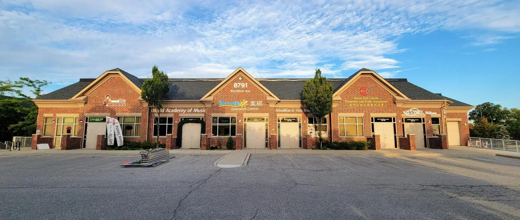 A single-story brick building hosts several businesses, including a music academy and a BBQ restaurant. With trees out front and a clear sky above, the facility showcases its fresh commercial shingle roofing. The parking lot is mostly empty, with a small stack of materials on the ground. Dominion Roofing | BRAMPTON | BURLINGTON | ETOBICOKE | MARKHAM | MISSISSAUGA | NORTH YORK | OAKVILLE | RICHMOND HILL | Scarborough | TORONTO | VAUGHAN | dominionroofing.com | Phone Number: 416-789-0601