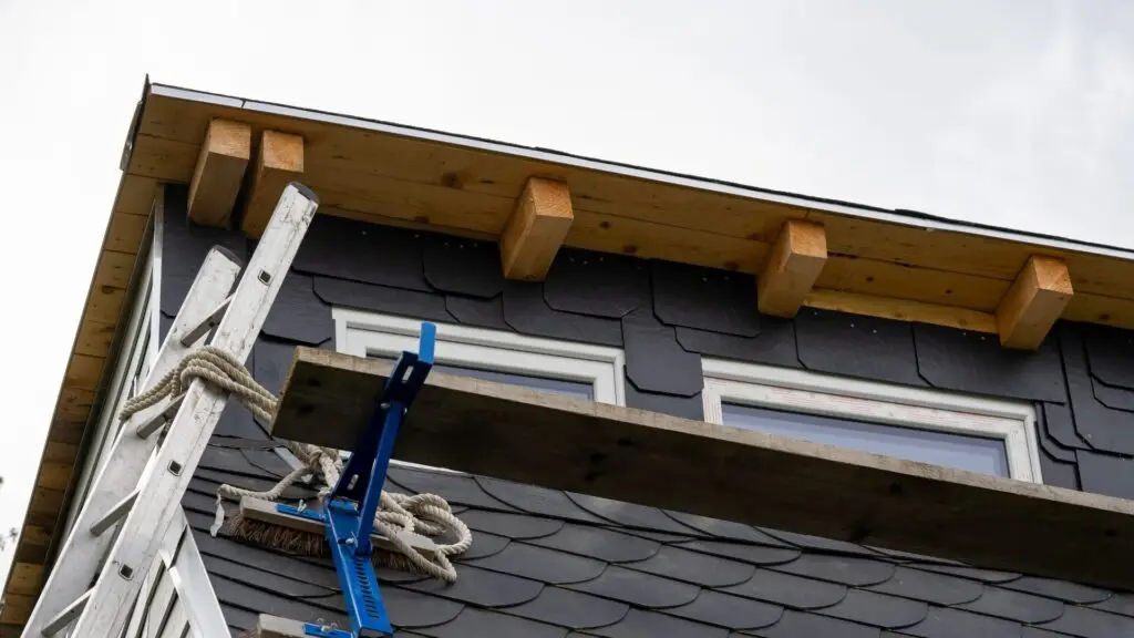 A section of a buildings roof is under construction. The structure features wooden supports beneath a black-tiled roof. A ladder is propped against the building, and scaffolding with rope is visible. Two windows are present below the roof edge. Dominion Roofing | BRAMPTON | BURLINGTON | ETOBICOKE | MARKHAM | MISSISSAUGA | NORTH YORK | OAKVILLE | RICHMOND HILL | Scarborough | TORONTO | VAUGHAN | dominionroofing.com | Phone Number: 416-789-0601
