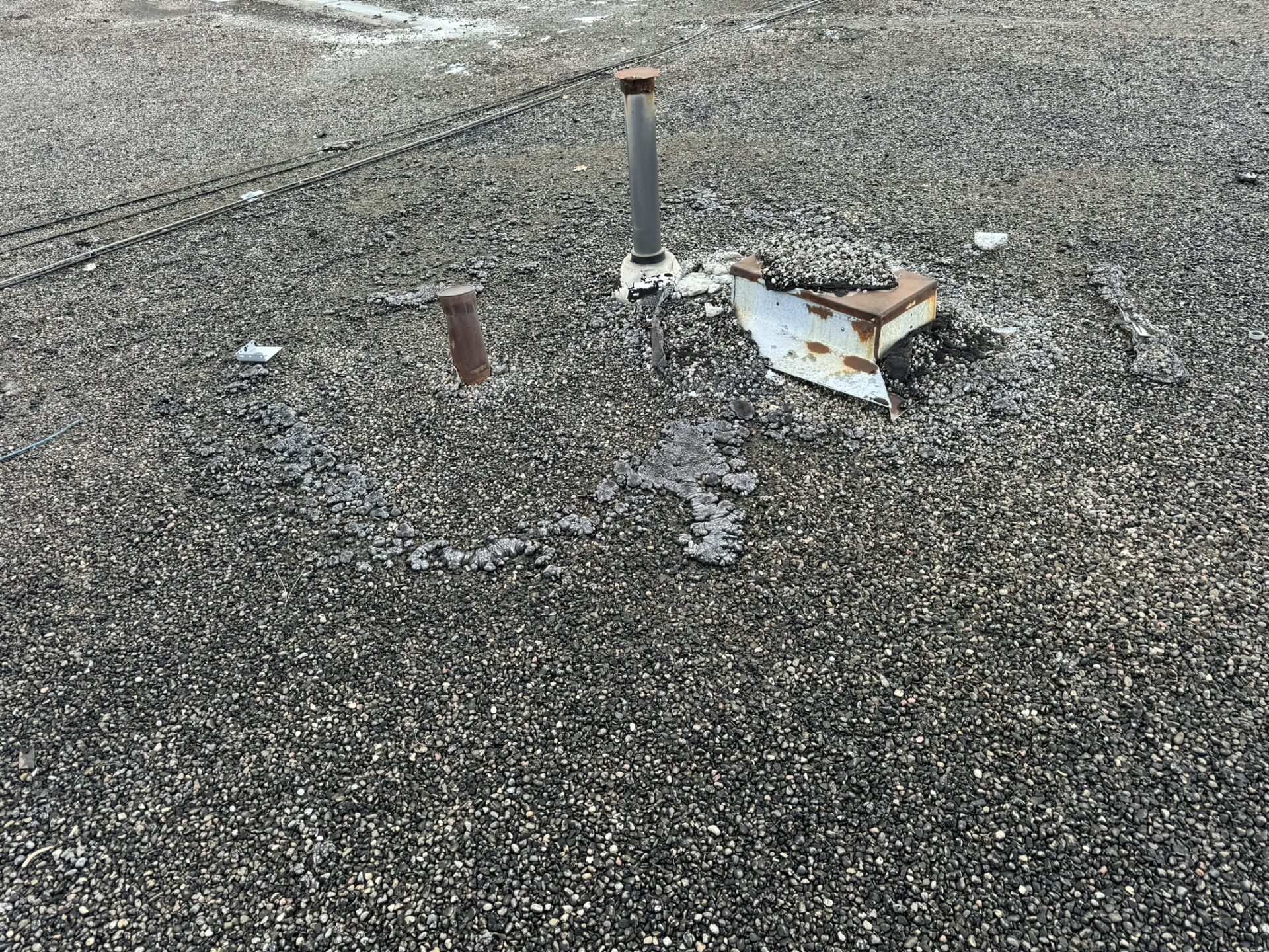 A flat, gravel-covered rooftop with two metal pipes protruding and a rusted metal vent lying on its side suggests potential roof repair needs. Patches of pebbles have gathered around the pipes and vent, forming a slightly raised area. Dominion Roofing | BRAMPTON | BURLINGTON | ETOBICOKE | MARKHAM | MISSISSAUGA | NORTH YORK | OAKVILLE | RICHMOND HILL | Scarborough | TORONTO | VAUGHAN | dominionroofing.com | Phone Number: 416-789-0601