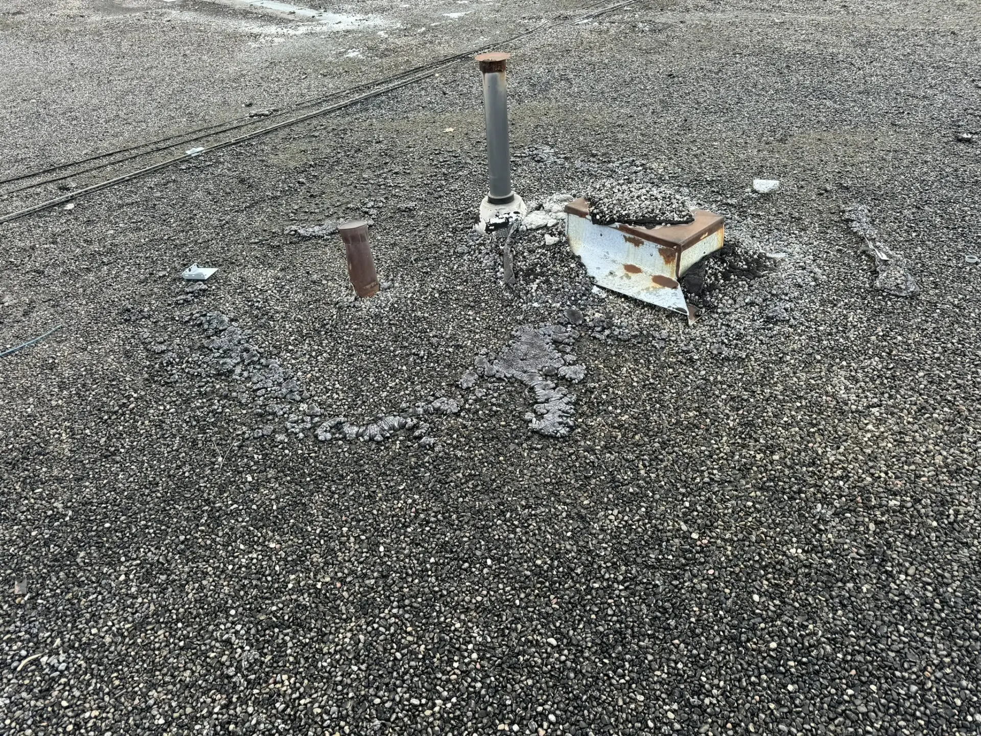 A flat, gravel-covered rooftop with two metal pipes protruding and a rusted metal vent lying on its side suggests potential roof repair needs. Patches of pebbles have gathered around the pipes and vent, forming a slightly raised area. Dominion Roofing | BRAMPTON | BURLINGTON | ETOBICOKE | MARKHAM | MISSISSAUGA | NORTH YORK | OAKVILLE | RICHMOND HILL | Scarborough | TORONTO | VAUGHAN | dominionroofing.com | Phone Number: 416-789-0601